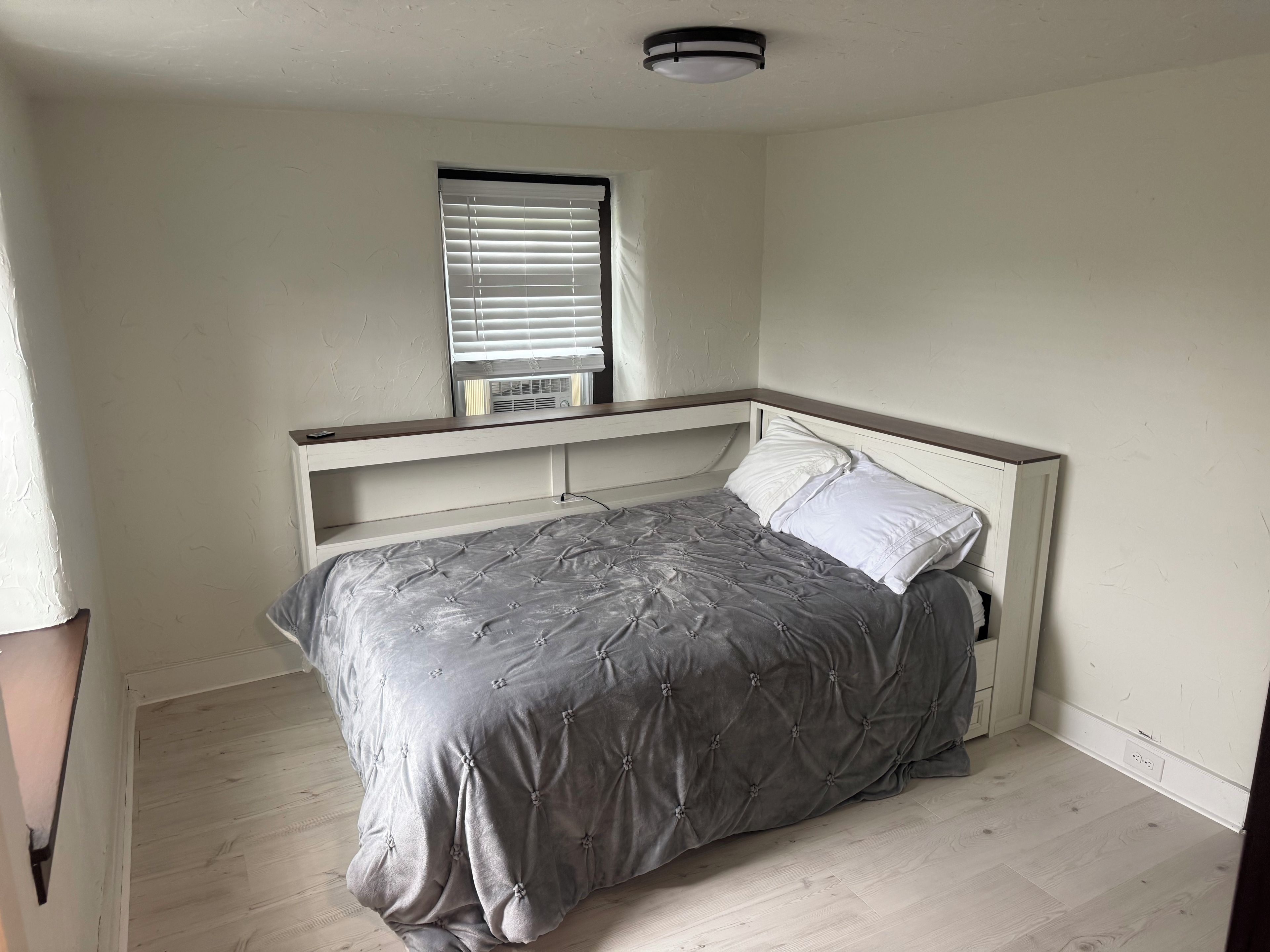 Een slaapkamer met een bed, een raam met zonwering, een plafondlamp en een wandairconditioner.