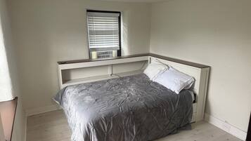 Een slaapkamer met een bed, een raam met zonwering, een plafondlamp en een wandairconditioner.