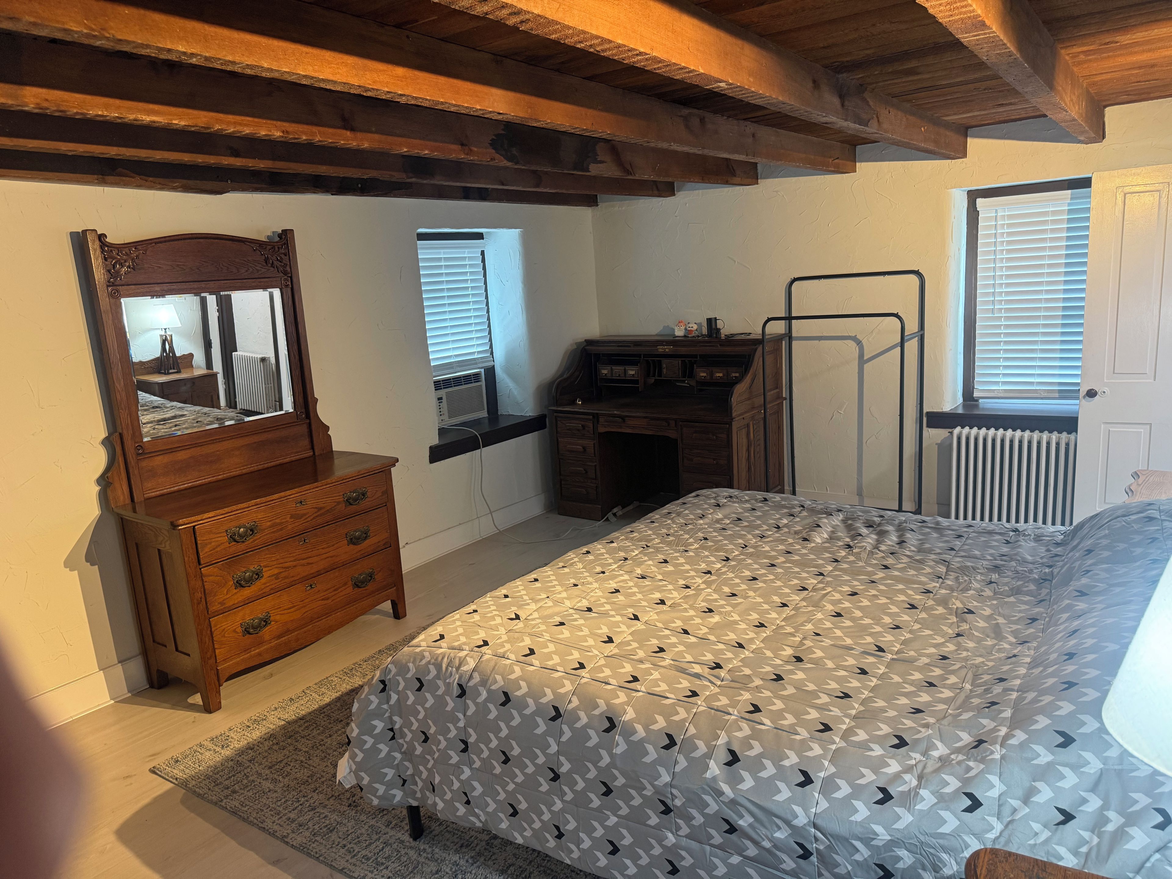 Een slaapkamer met een bed, een kaptafel, een spiegel, een bureau, een radiator en ramen.