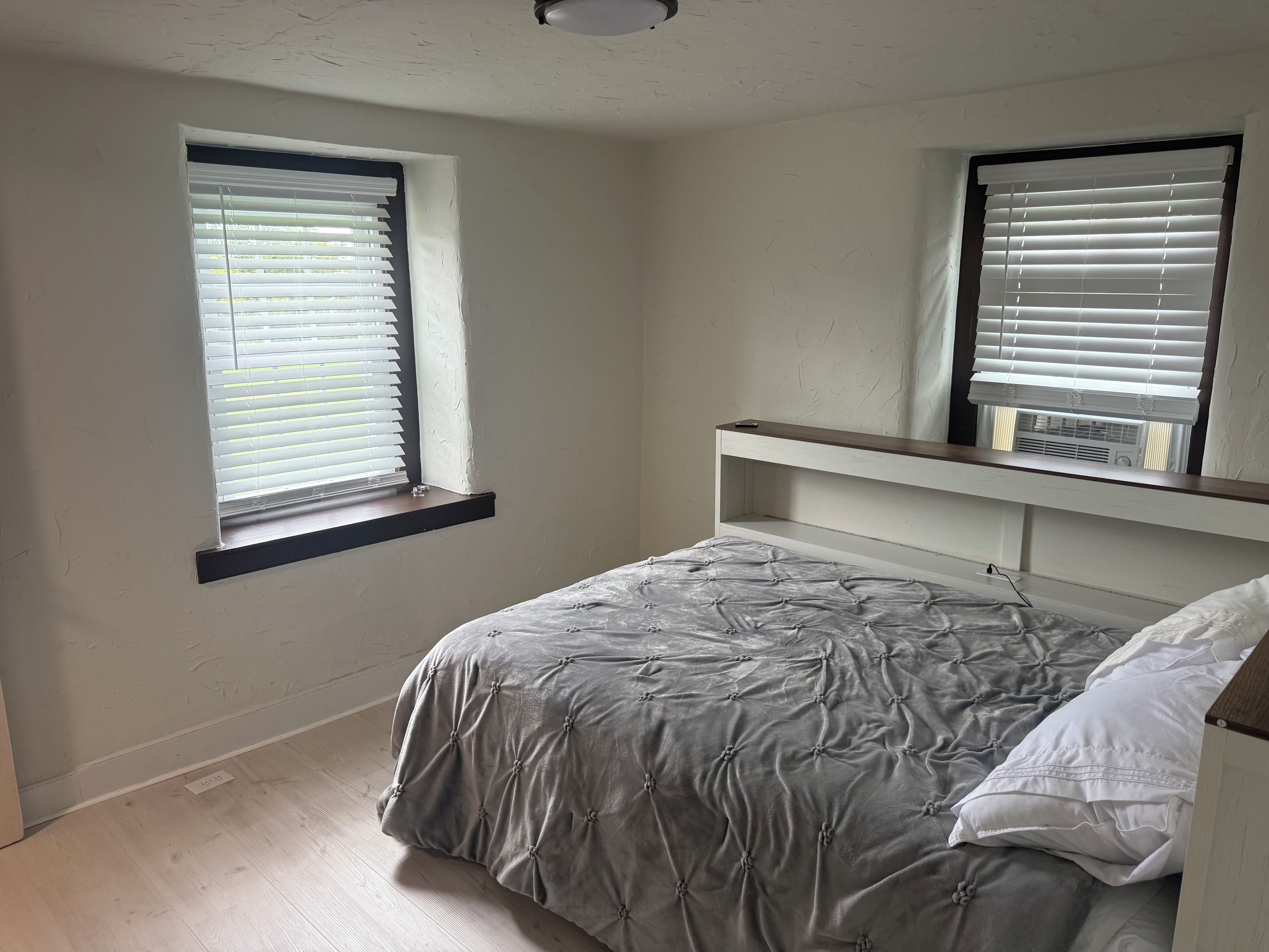 Une chambre à coucher avec un lit, deux fenêtres équipées de stores, un luminaire au plafond et une étagère intégrée.