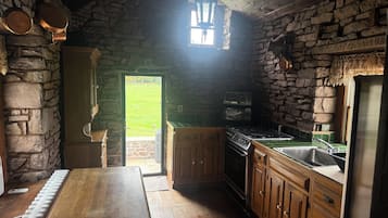 A rustic kitchen with stone walls, wooden cabinets, a built-in oven, and a window overlooking a grassy area.