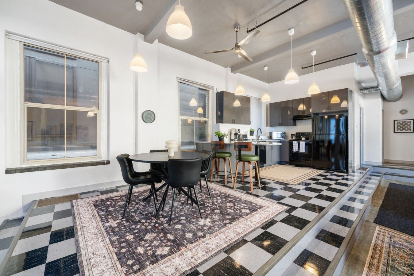 Una cocina moderna con piso a cuadros blanco y negro, iluminación colgante y un comedor con alfombra estampada.