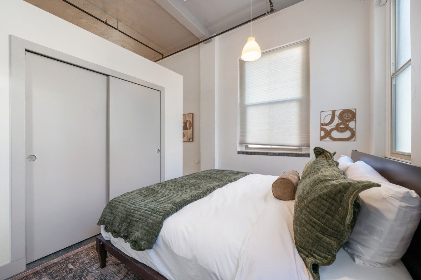 Une chambre à coucher avec un lit, une fenêtre avec des stores, un luminaire suspendu et une porte.