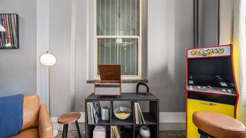 Um quarto com janela, estante de livros, máquina de arcade Pac-Man, luminária e poltrona de couro.