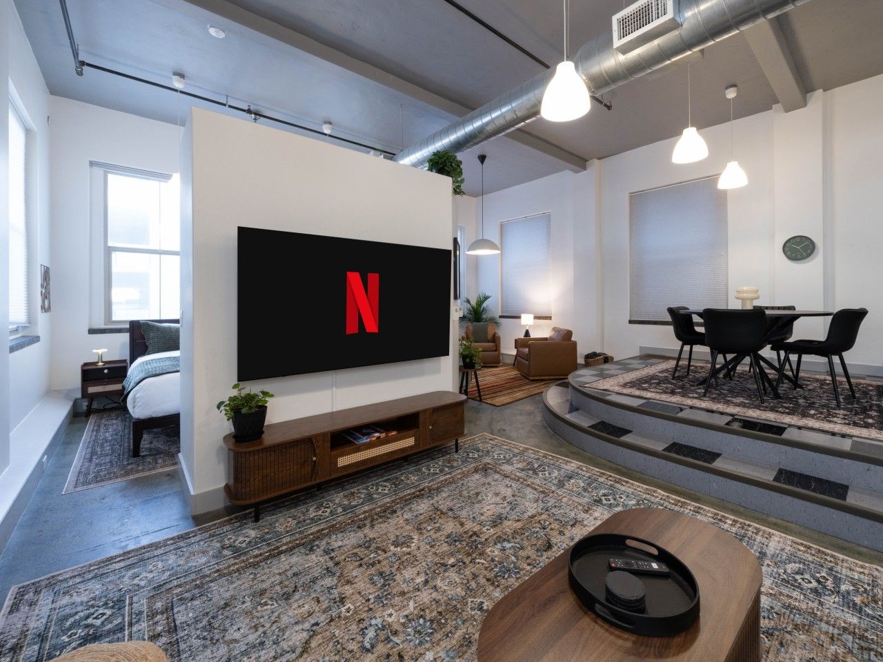 A modern living room with a large flat-screen TV mounted on the wall, a seating area with a sofa and chairs, a coffee table, and a patterned rug.