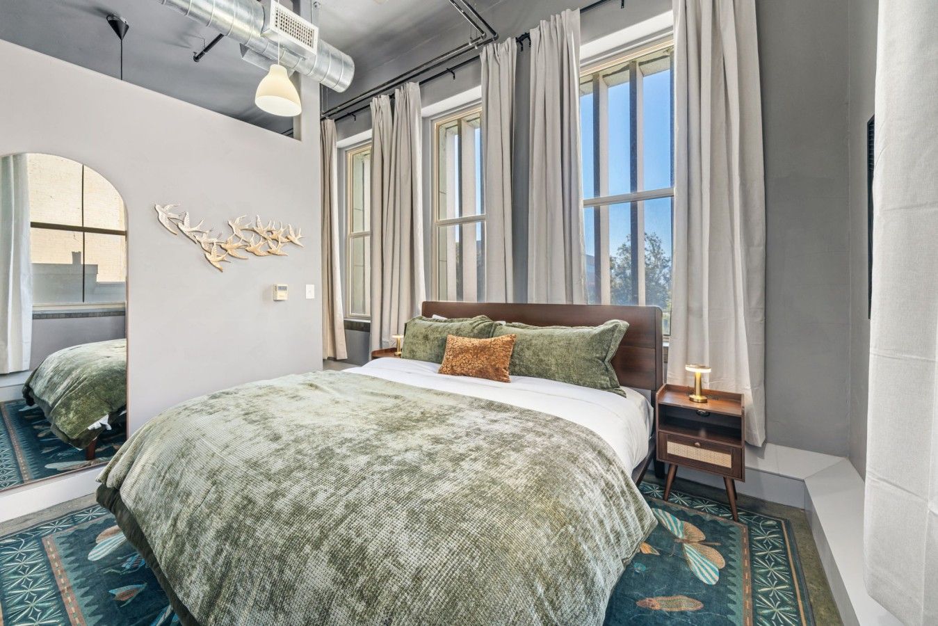 Quarto com cama grande, mesa de cabeceira, espelho e janela com cortinas.