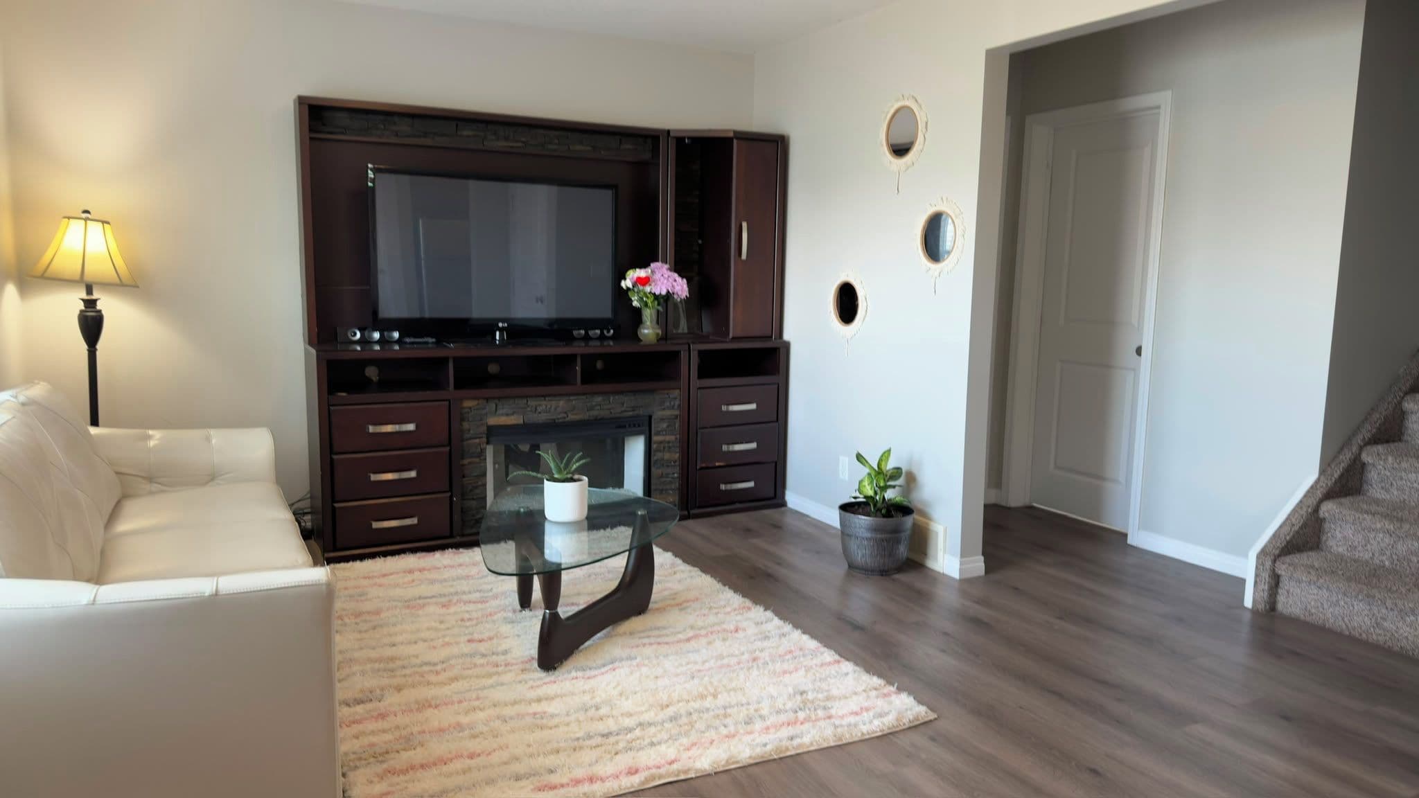 Una sala de estar con un televisor de pantalla plana, un mueble de madera oscura para equipo multimedia, una mesa de centro de vidrio, un sofá blanco y una escalera que conduce a otro nivel.