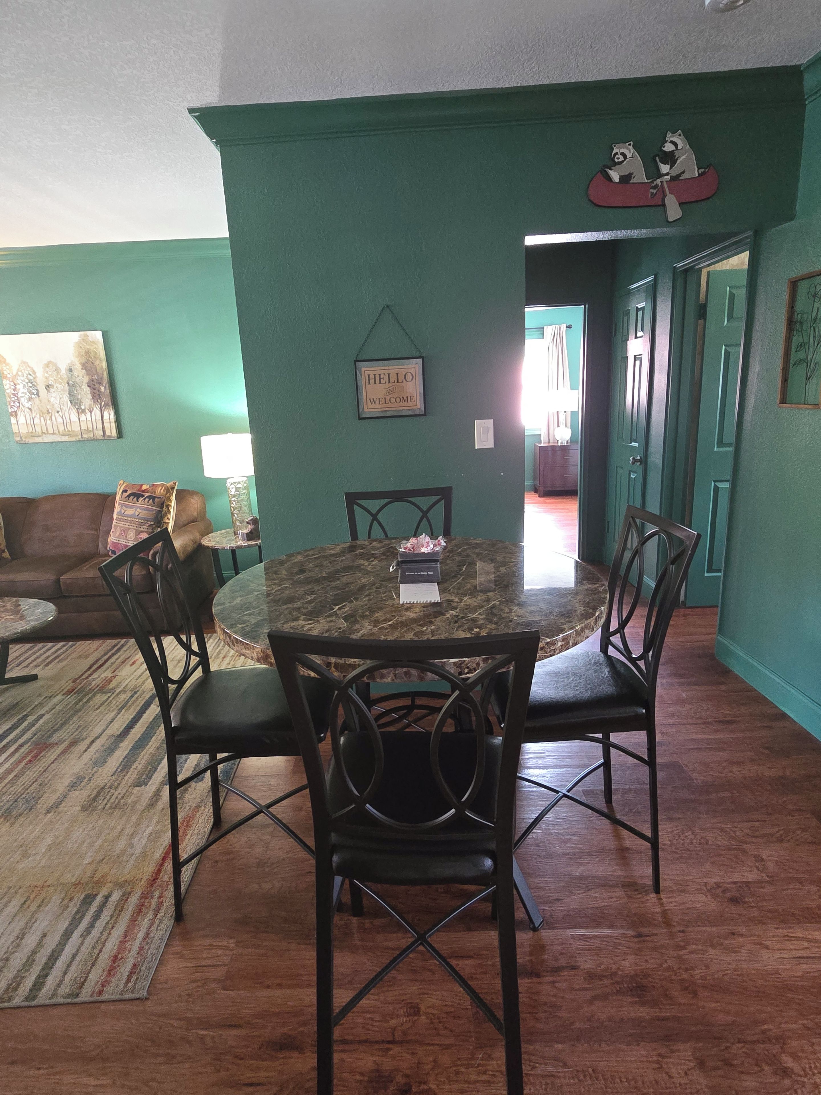 A dining area with a round table, four chairs, a green wall, and a 'HELLO WELCOME' sign.