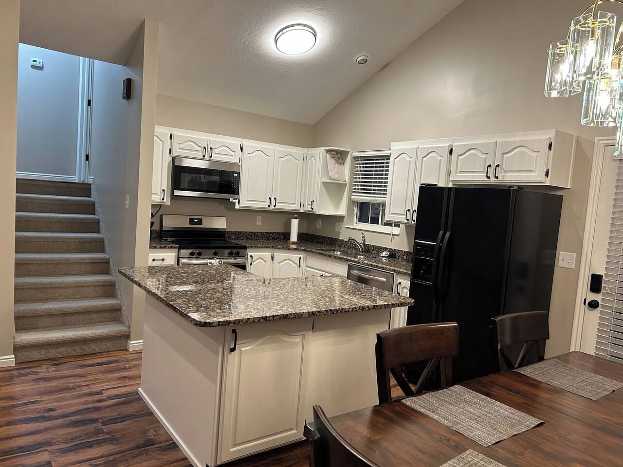 Una cocina con armarios blancos, encimera de granito, refrigerador negro y una zona de comedor con sillas y mesa de madera.