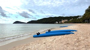 A beach with kayaks lined up on the sand, a calm sea, and a building with a tower in the background.