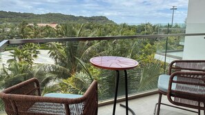 Varanda com cadeiras de vime e uma mesa pequena, com vista para uma paisagem tropical com palmeiras e uma colina ao longe.