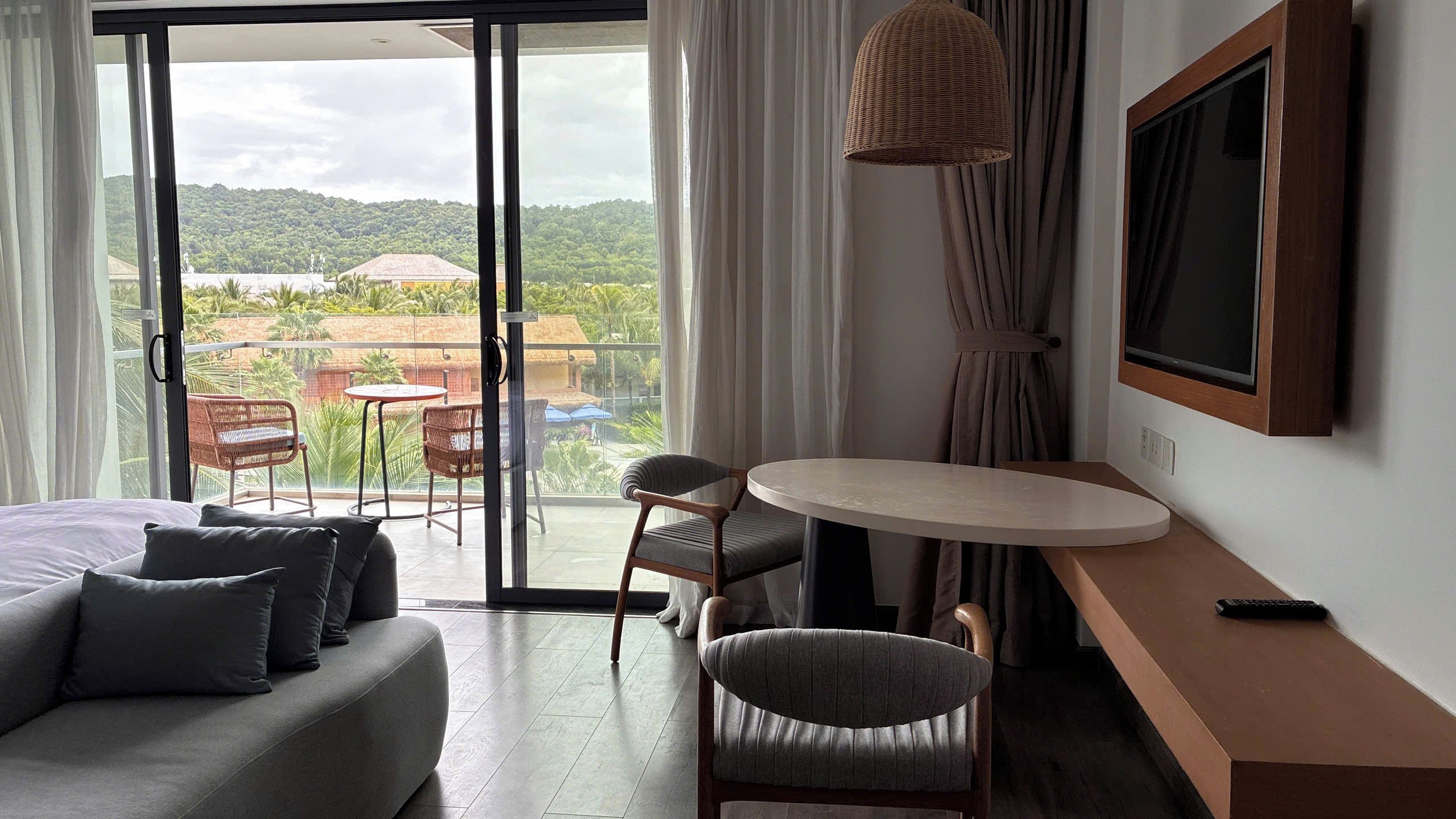 A hotel room with a bed, a round table, two chairs, a TV, and a balcony with a view of greenery.