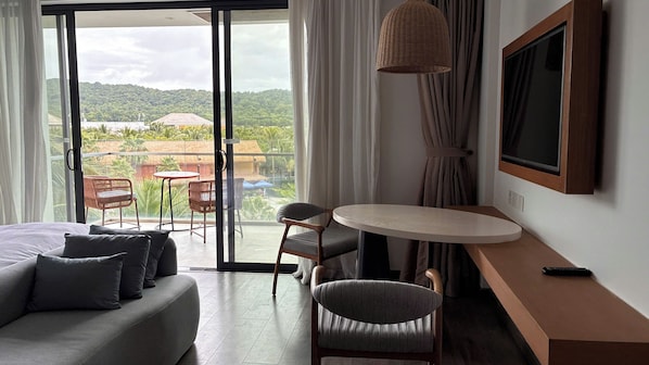 A hotel room with a bed, a round table, two chairs, a TV, and a balcony with a view of greenery. - DIAMOND SUITE KEM BEACH (Phu Quoc)