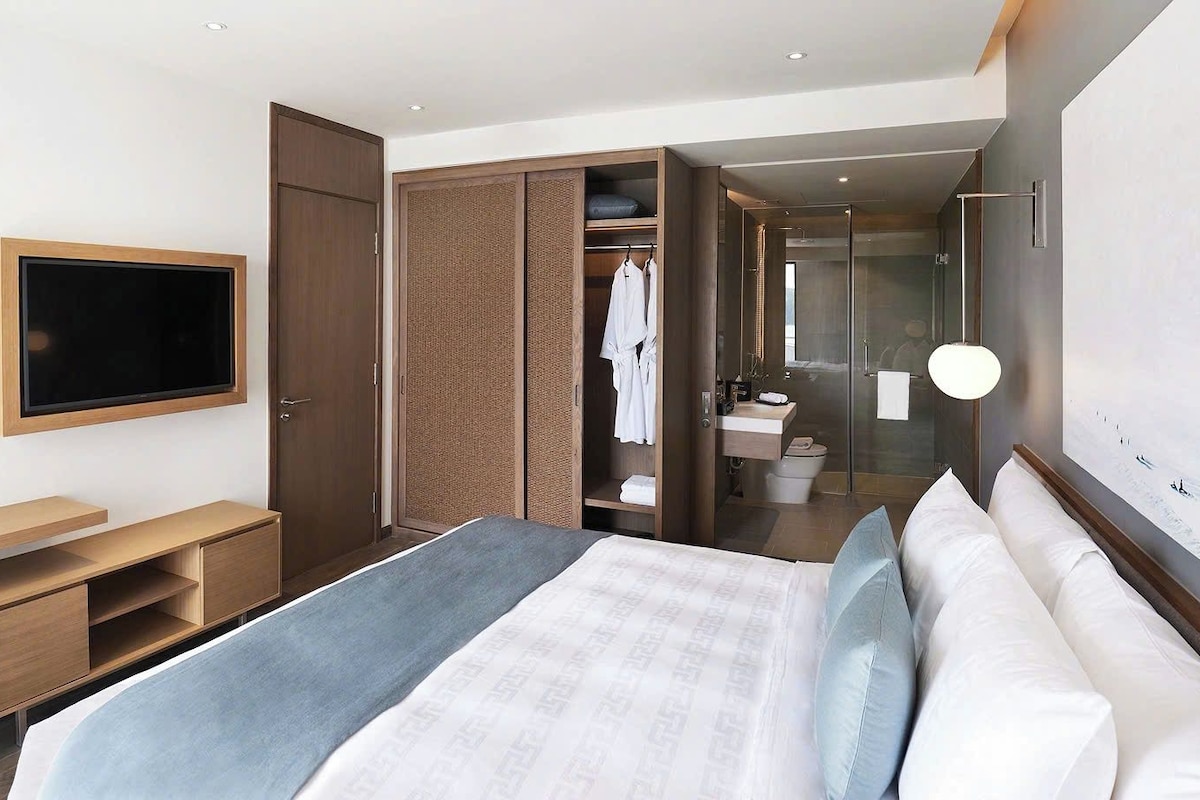 A modern hotel room with a bed, a TV, a desk, a wardrobe, and a bathroom visible through an open door.