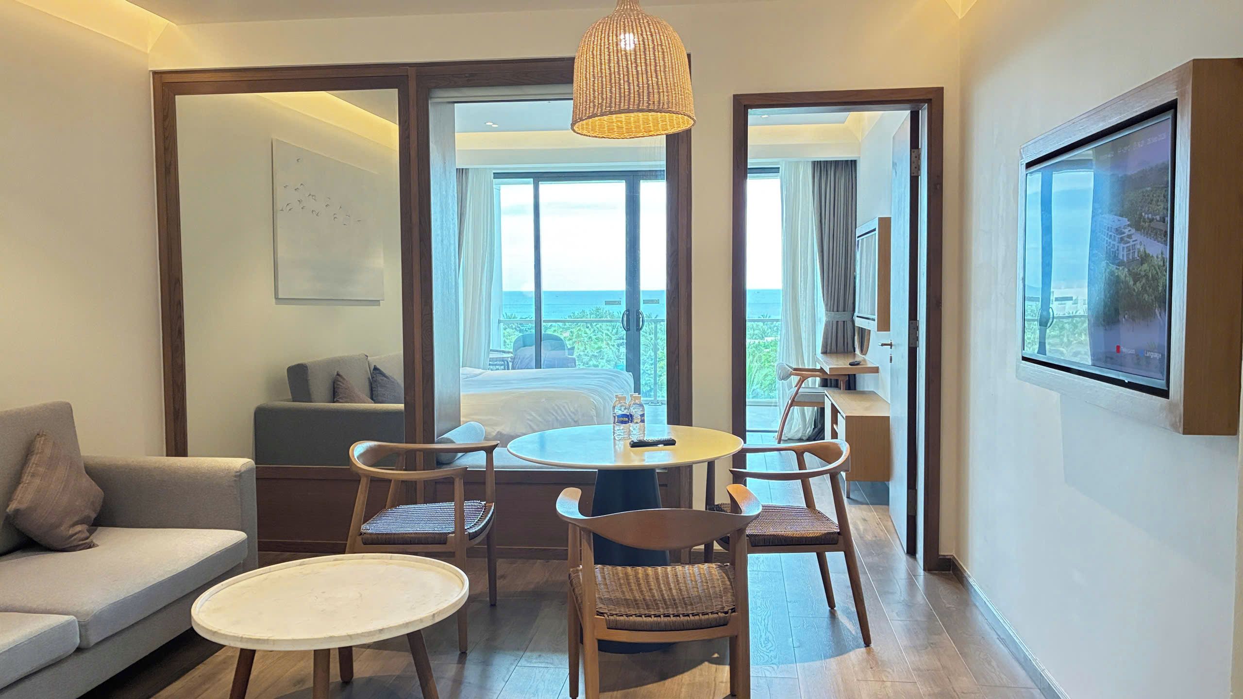 A hotel room with a sofa, a round dining table with chairs, a large window with a view of the sea, and a painting on the wall.