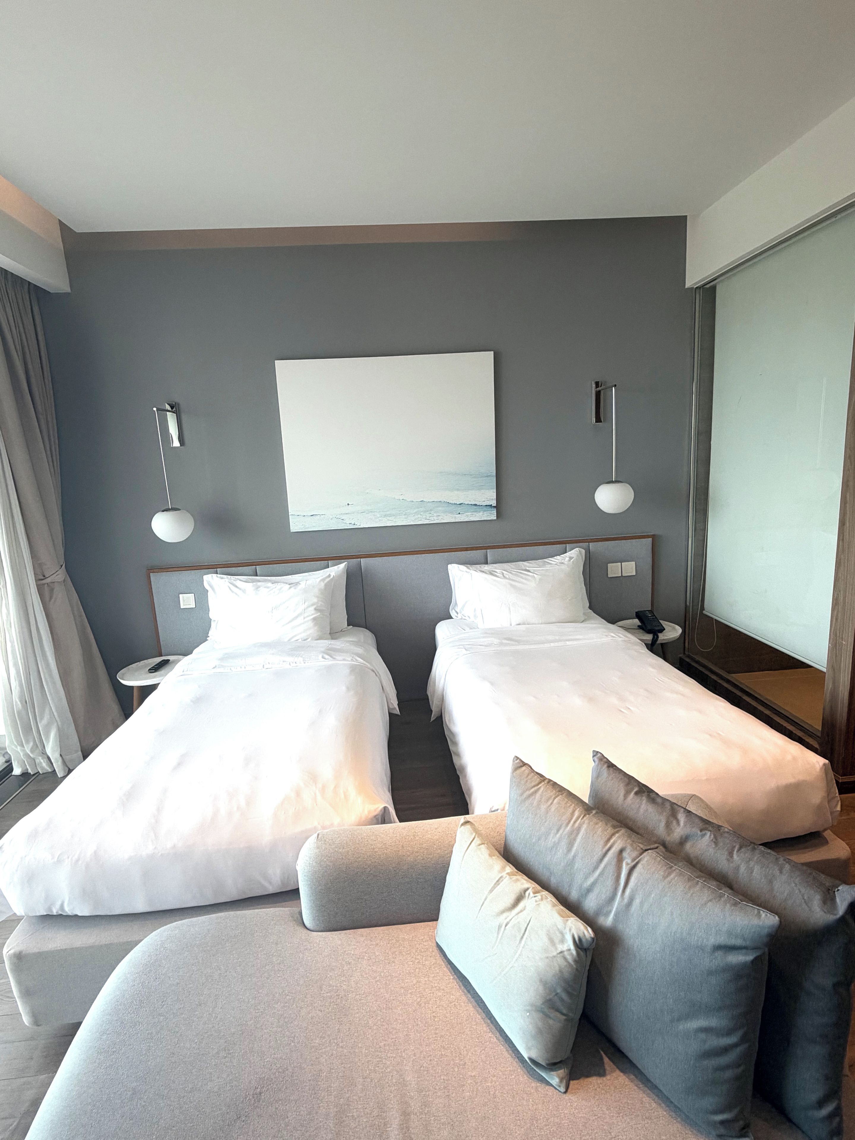 Habitación de hotel con dos camas individuales, un sofá, un ventanal grande y un cuadro en la pared.