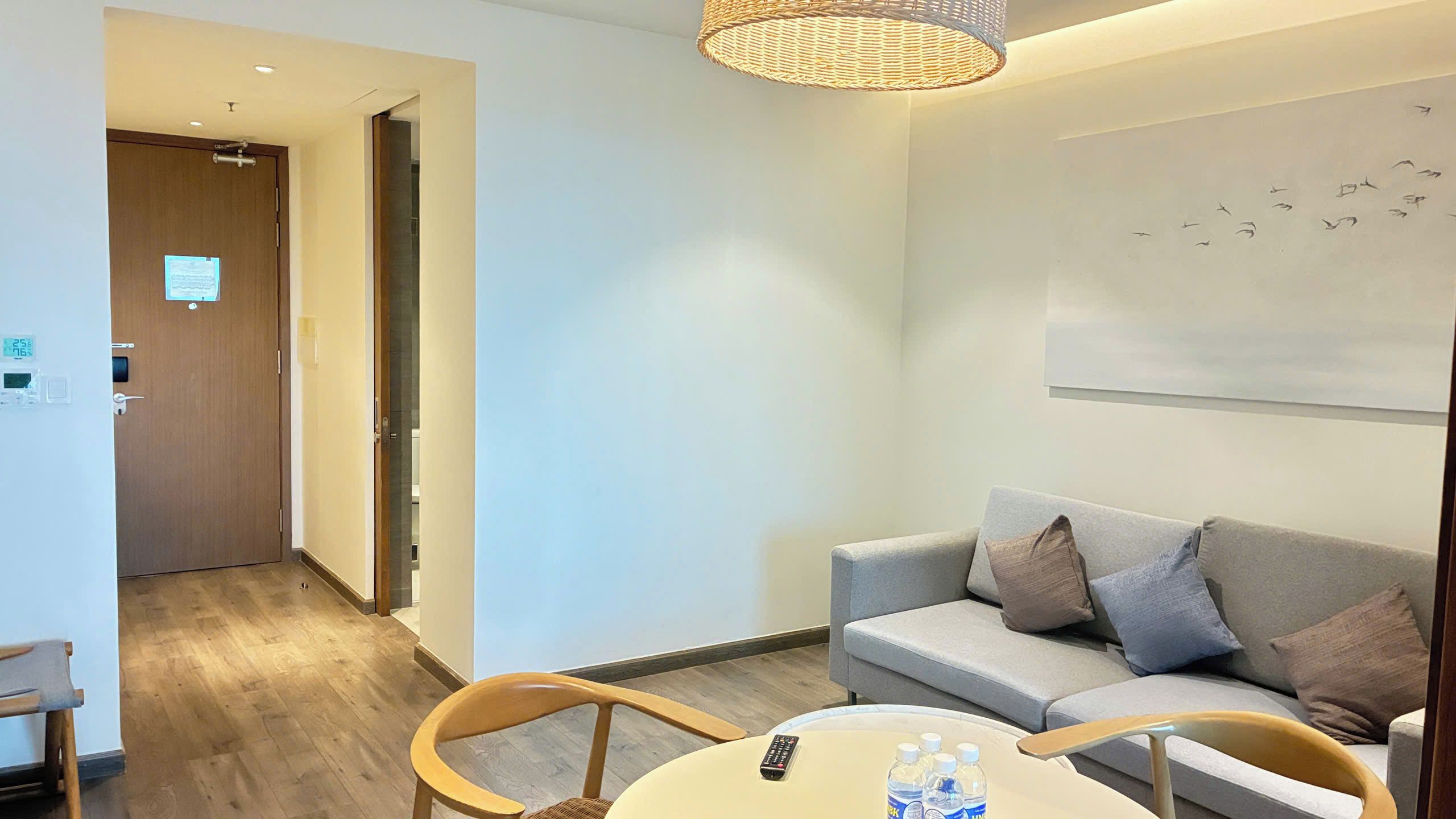 A modern hotel room with a grey sofa, a round table with a remote control, and a bottle of water.