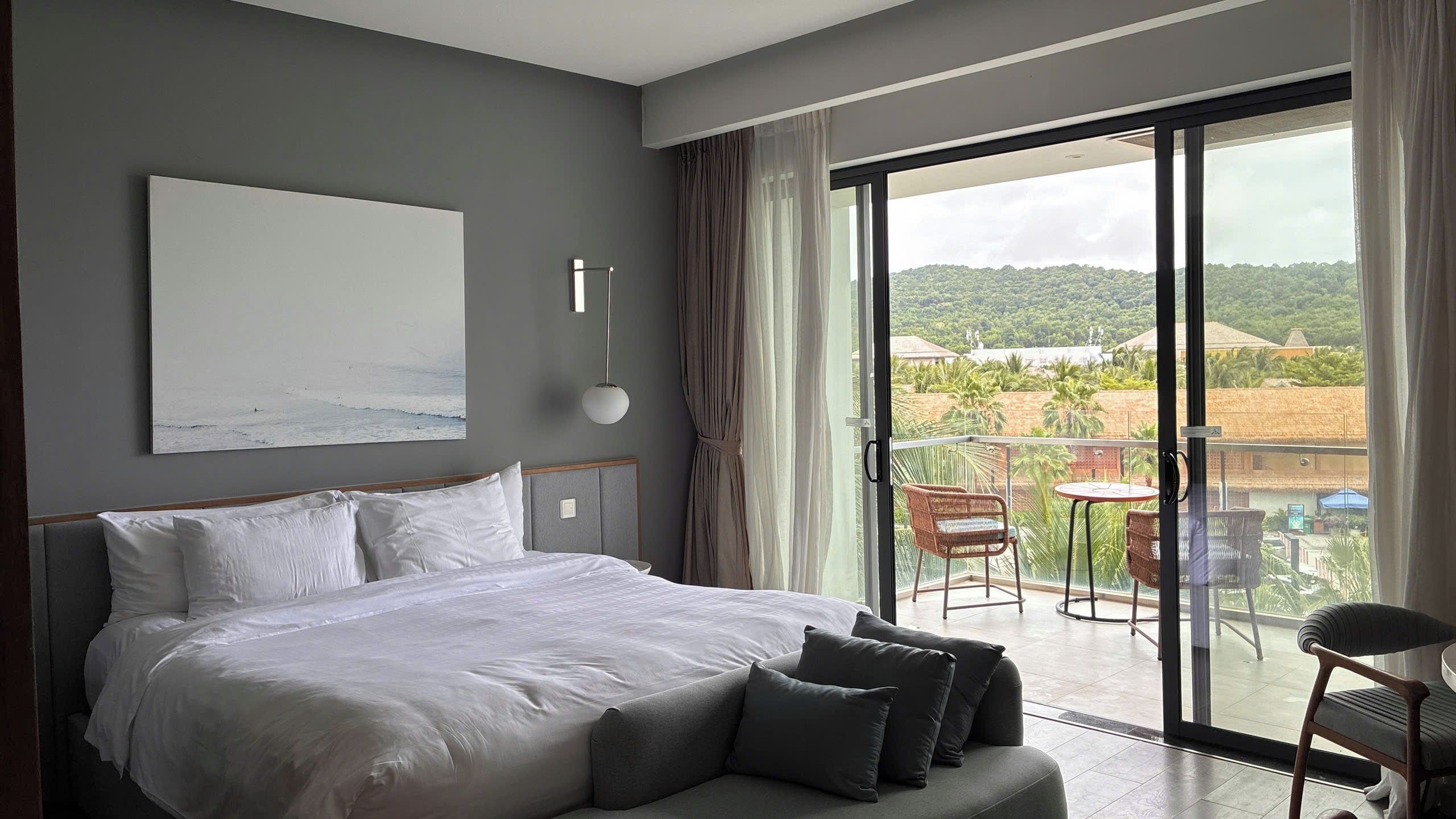A bedroom with a large bed, a view of the outdoors, and a balcony with chairs.