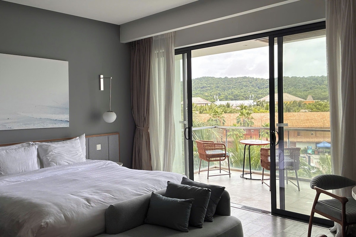 A bedroom with a large bed, a view of the outdoors, and a balcony with chairs.