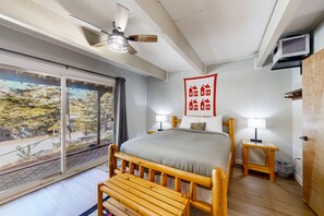 A bedroom with a large bed, wooden headboard, two bedside tables with lamps, a ceiling fan, and a view of trees and buildings through a sliding door. - Winhall townhome with ski mudroom and Bromley slope views from the balcony (Winhall)