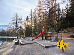 A playground with a red slide and climbing structures, surrounded by trees and a lake, with mountains in the background. - Chalet in Praz-de-Fort im Val Ferret by Interhome (Champex)