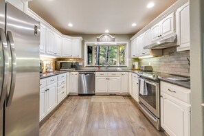 A kitchen with white cabinets, stainless steel appliances, and a view of trees through a window. - 8 Quelah - Sunriver Escape w/ 10 SHARC Passes, Pool Table, Deck (Sunriver)