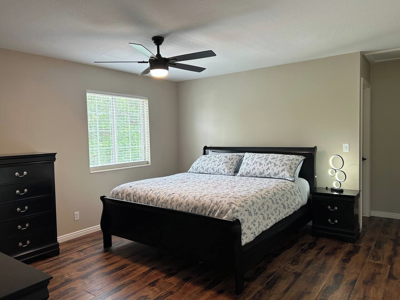 A bedroom with a bed, a dresser, a nightstand with a lamp, a ceiling fan, and a window with blinds.