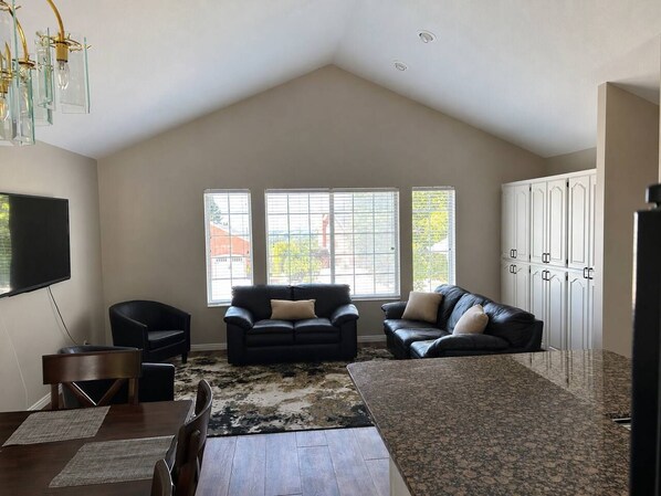 A spacious living room with a high ceiling, a large window, a flat-screen TV, two leather sofas, a dining table, and a kitchen area.