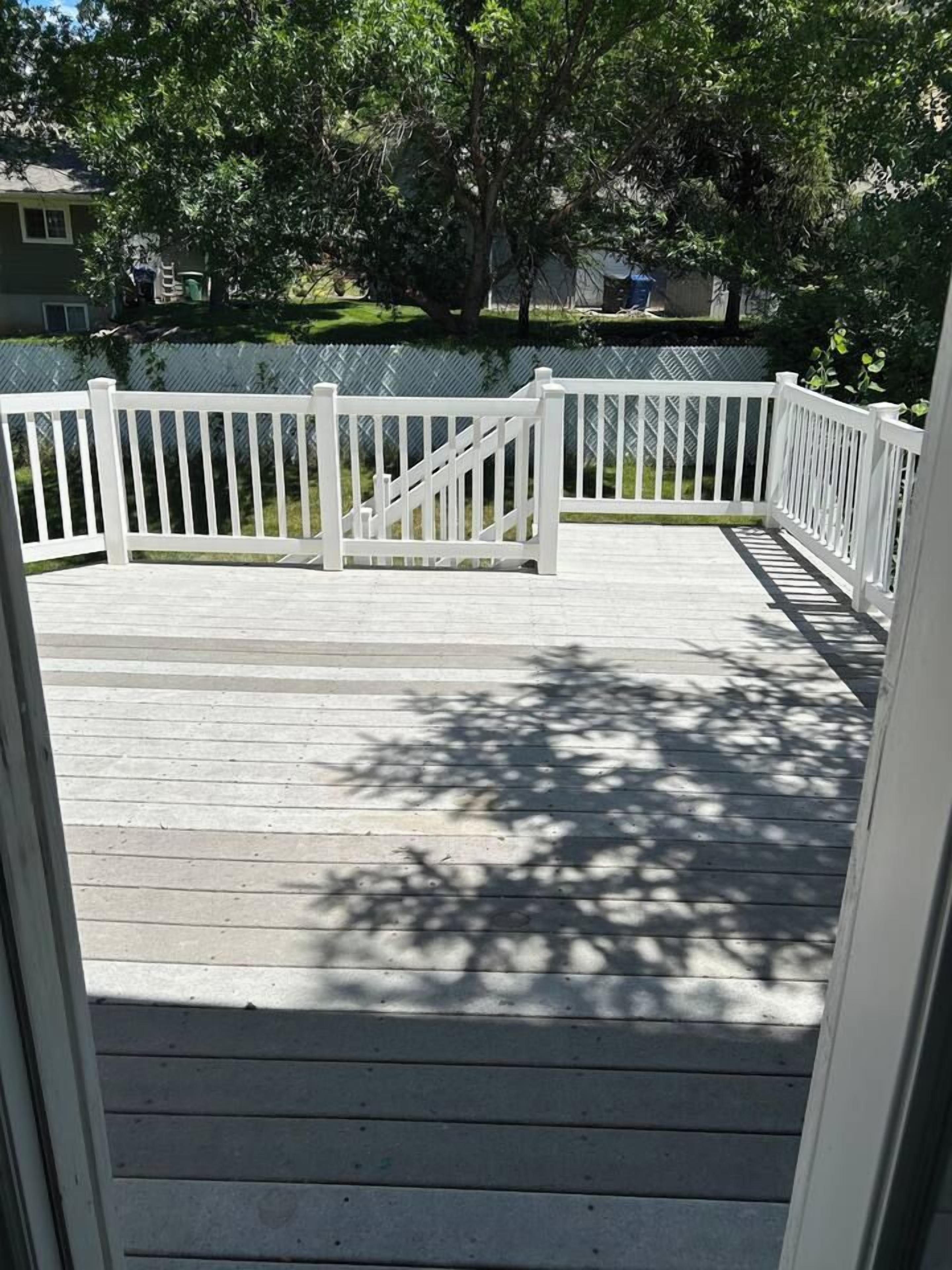 A wooden deck with white railings, surrounded by trees and a fence.
