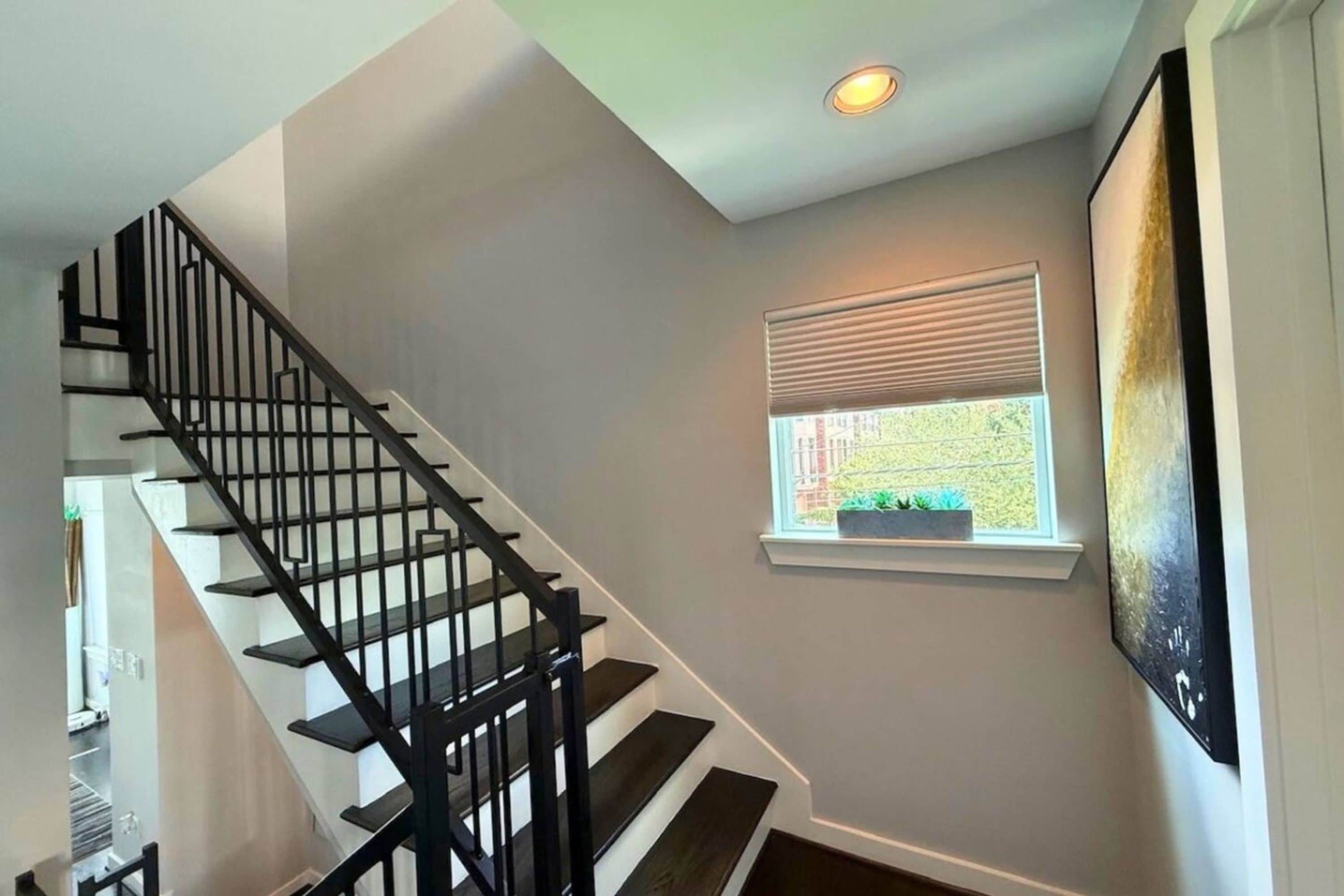 Un escalier avec des rampes noires menant à un palier, une fenêtre avec des stores et un spot encastré au plafond.