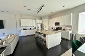 Uma cozinha moderna com armários brancos, uma ilha central com banquetas, eletrodomésticos de aço inoxidável e uma TV de tela plana na parede.