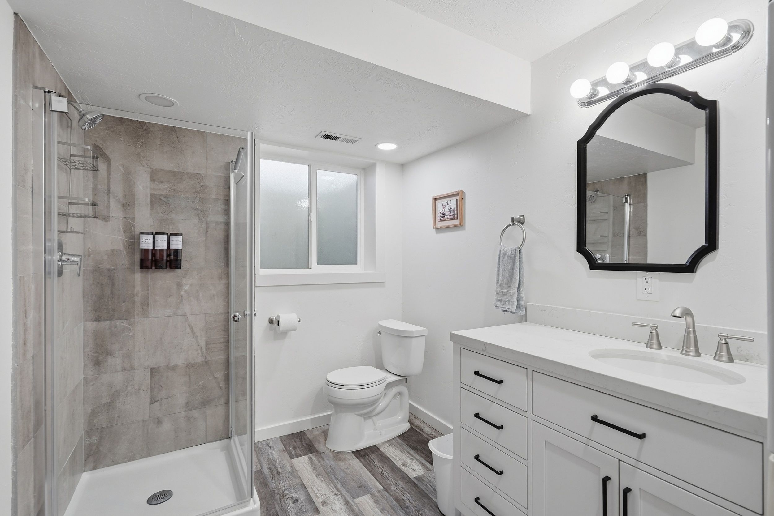 A bathroom with a shower, toilet, vanity, and a mirror with lights.