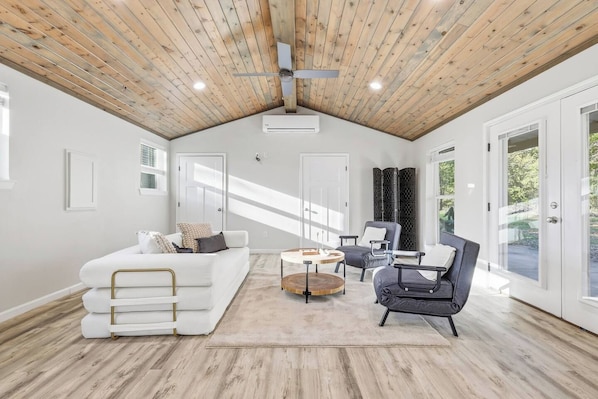 A spacious room with a sloped ceiling, a white sofa, a round coffee table, a black armchair, and a large window providing natural light.