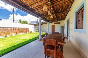 A covered patio with a long wooden table and chairs, a tiled floor, and a view of a green lawn. - House with a pool 350 m from Peroba Beach (Maragogi)