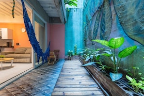A narrow wooden pathway leading to a seating area with a sofa and a table, surrounded by potted plants and a blue hanging textile. - Utopia S East Quiet Loft Wi-fi Self Check-in (Medellín)