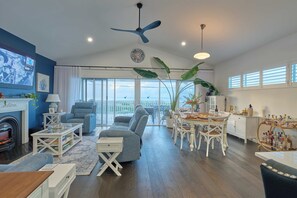 A spacious living room with a fireplace, a large sofa, a dining area with white chairs, and a view of the sea through large windows. - Blu Bay View—Stroll Down to the Bay at Golden Hour (Geographe)