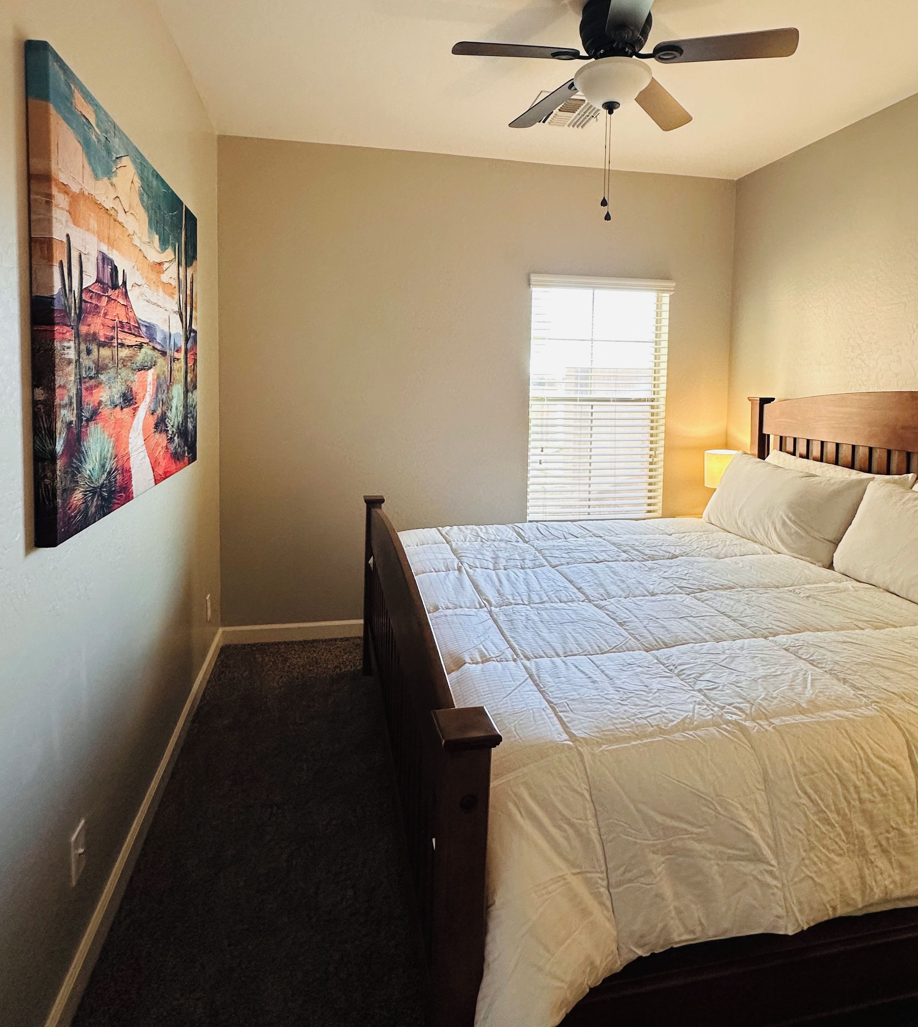 A bedroom with a bed, a ceiling fan, a window with blinds, and a painting on the wall.