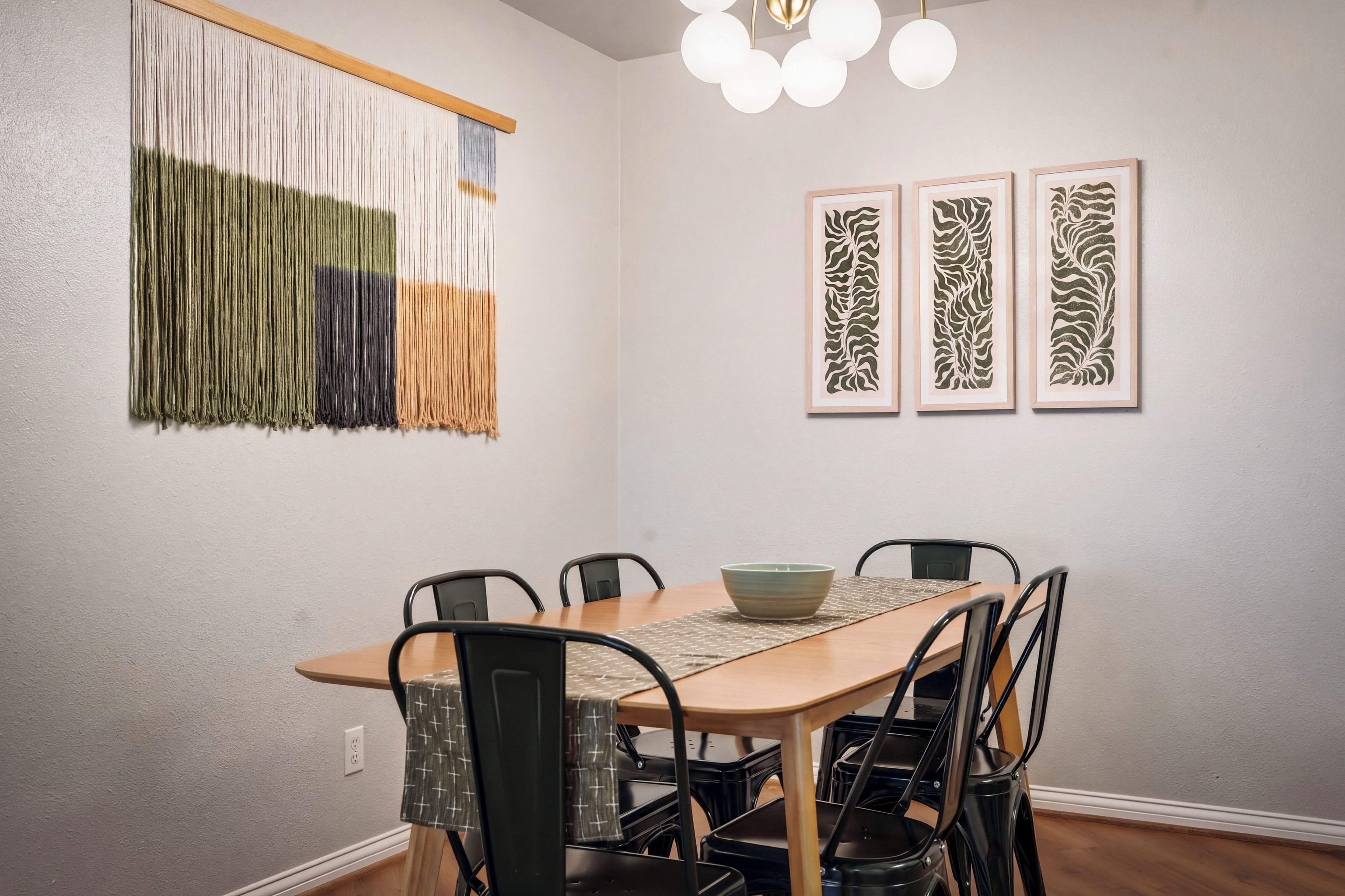 Un comedor con una mesa de madera, sillas negras, un tapiz tejido y tres cuadros enmarcados.