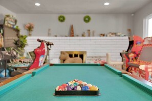 A pool table with a ball rack in the foreground, a red flamingo decoration, and a fireplace with a wreath on the wall. - Travelers Dream|Monthly Stays|Strip in the Horizon (Las Vegas)