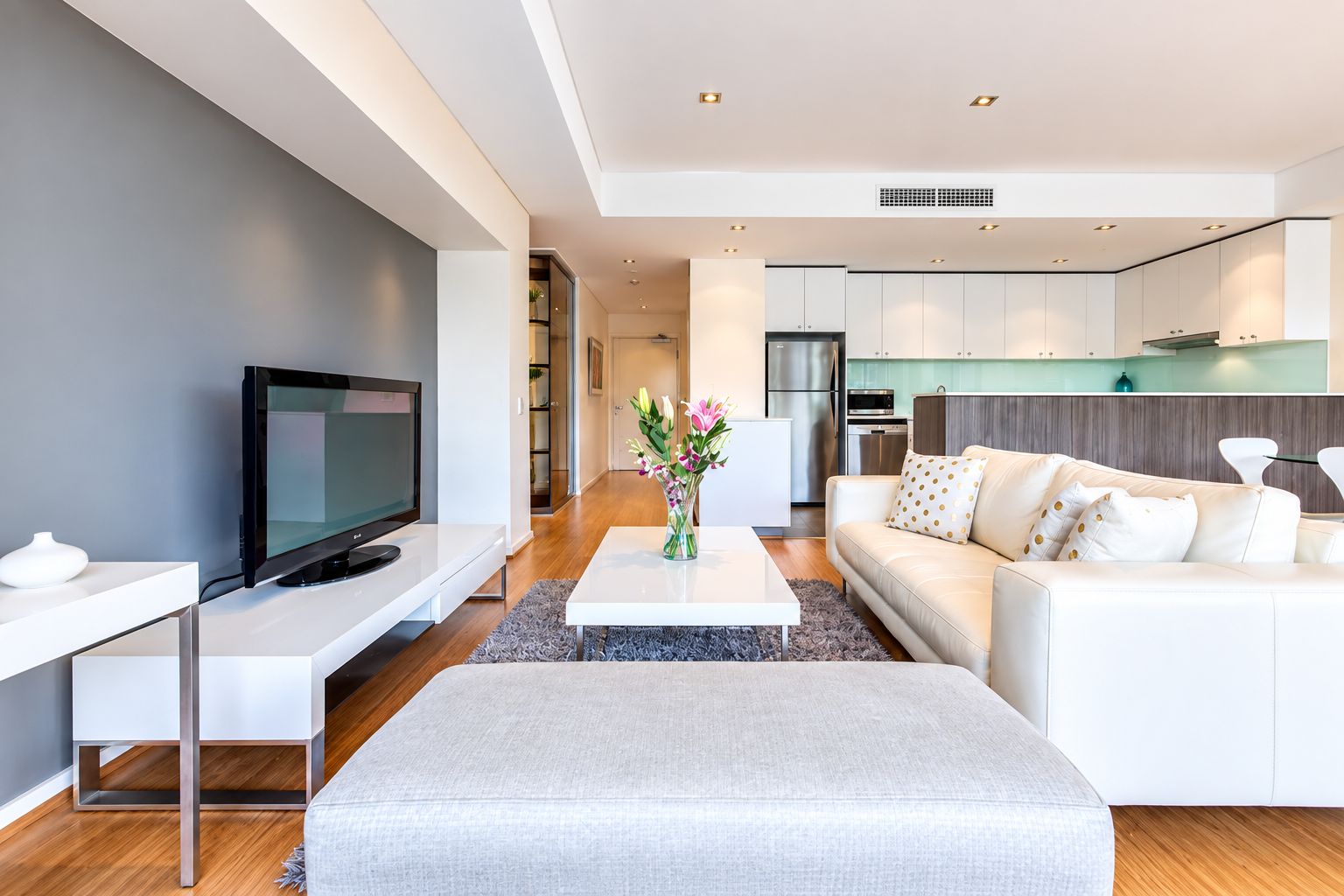A modern living room with a flat-screen TV, a white sofa, and a coffee table with a vase of flowers.