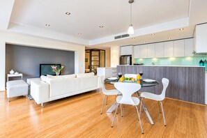 A modern kitchen with white cabinets, a dining area with a glass table and white chairs, and a living area with a white sofa and a flat-screen TV. - Central Freo 3BR Apartment, Pool, Gym & parking (Fremantle)