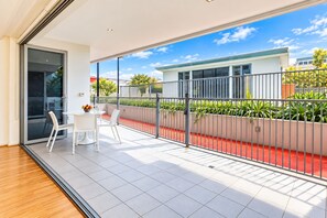 Ein moderner Balkon mit Glastür, weißen Stühlen, einem kleinen Tisch und Blick auf ein Gebäude über einem mit roten Ziegeln bedeckten Bereich.