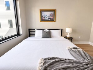 Ein Hotelzimmer mit einem großen Bett, einem Bild an der Wand, einer Lampe und einem Fenster mit Blick auf Gebäude.