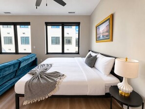 Una habitación de hotel con una cama, una mesita de noche con lámpara, un sofá azul y ventanas amplias.