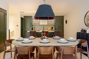 Comedor con mesa redonda dispuesta para seis personas, rodeada de sillas de madera, una lámpara colgante y una cocina al fondo.