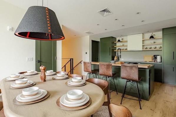 Un comedor moderno con una mesa redonda preparada para seis personas, una cocina con armarios verdes y una escalera que conduce a otro nivel.