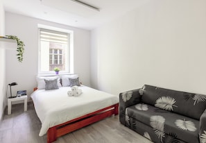 A bedroom with a bed, bedside table, lamp, and a window with a view. - Piłsudskiego City Center Apartment (Szczecin)