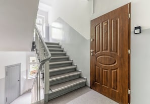 A staircase leading to a wooden door with a number plate, a metal handrail, and a small sign on the wall. - Piłsudskiego City Center Apartment (Szczecin)