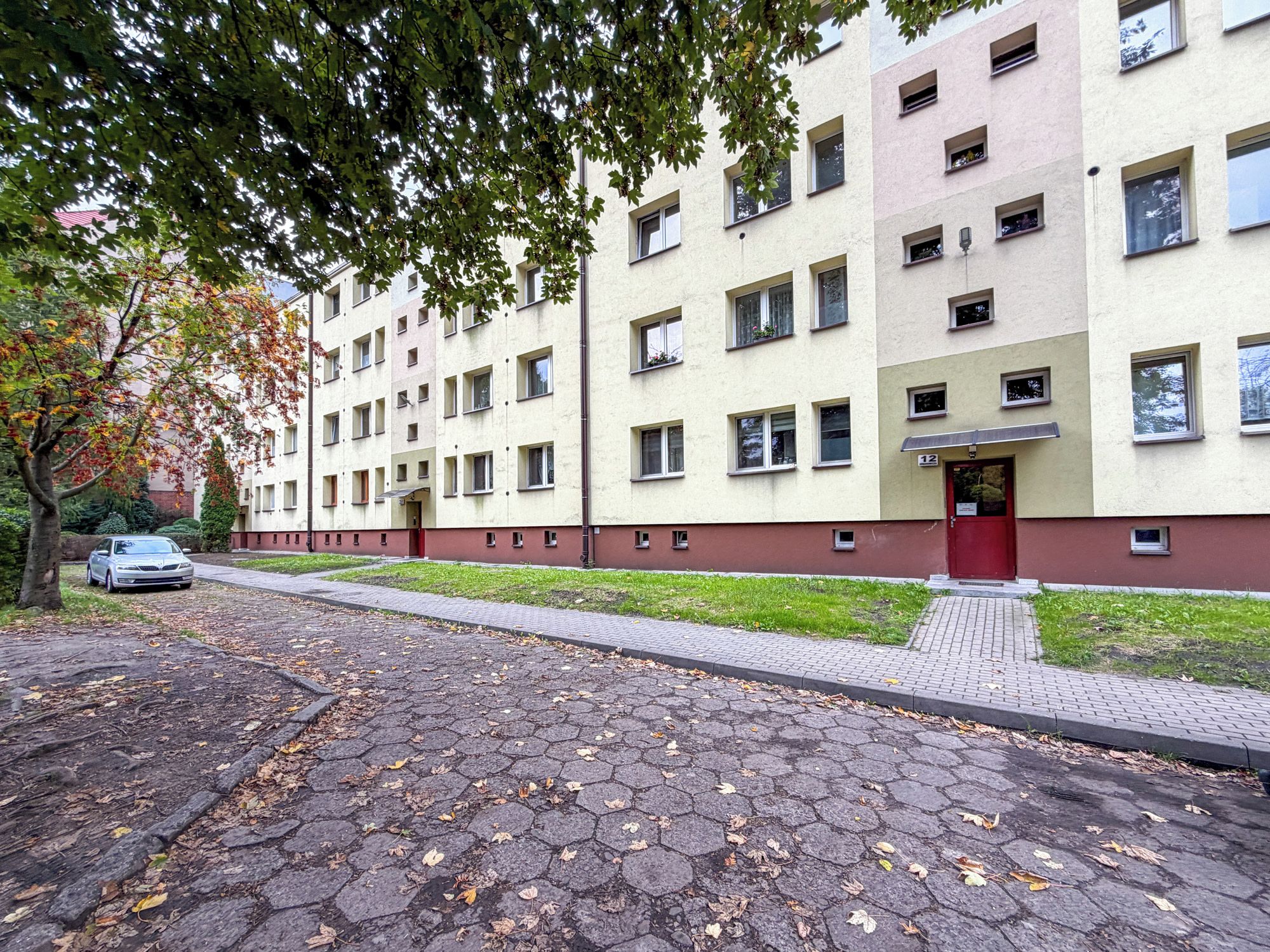 Un edificio residencial de varios pisos con una puerta roja, número 12, y un camino pavimentado cubierto de hojas caídas.