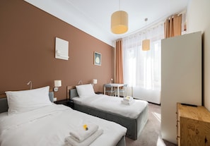 A hotel room with two beds, a desk, a chair, a window with curtains, and a wall-mounted light fixture.